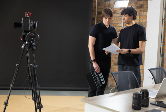Two adult men wearing black standing in studio by black backdrop tripod holding clapperboard script