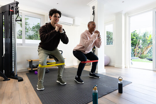 African American men wearing hoodies performing squats with yellow and red loop bands in home gym