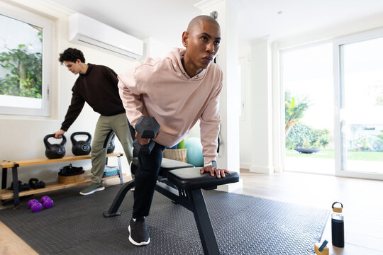 African male friends training in home gym, performing hex dumbbell row on bench