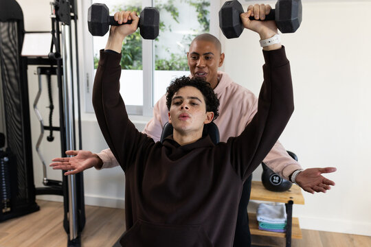African American male trainer spotting client pressing hex dumbbells on incline bench in gym