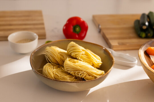 Ceramic bowl is holding four thin pasta nests on light countertop with red pepper and zucchinis