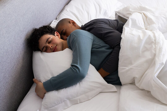 Two men spooning and resting in bedroom on bed with white pillows and grey headboard