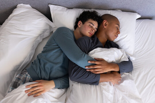 Two African American men cuddling and sleeping in bed wearing pajamas and white duvet pillows
