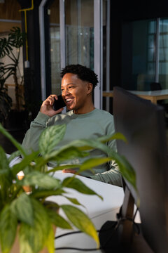 African American man talking on phone, sitting at desk in light green sweatshirt with potted plant