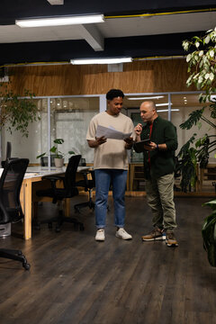 Diverse male coworkers wearing casual tee and jacket reviewing documents and tablet by glass wall