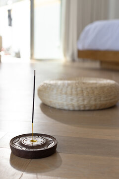 Single lit incense stick is releasing thin smoke from wooden holder on light floor near window