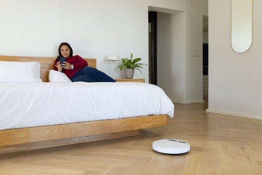 Adult Asian woman reclining on platform bed in bedroom, holding smartphone, robot vacuum operating