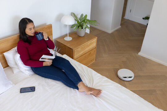 Female reclining on bed holding mug, reading open book, phone near, robot vacuum running