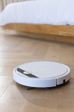 White robotic vacuum cleaner is sitting on light parquet floor in bedroom, near rumpled duvet