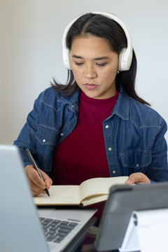 Asian woman wearing denim button-up, white headphones writing in notebook at laptop and tablet