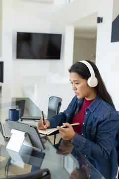 Woman wearing denim jacket and white headset writing in notebook at glass table using tablet stand