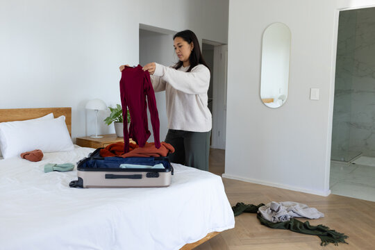 Asian female standing by bed holding maroon long-sleeve top and packing suitcase on bed