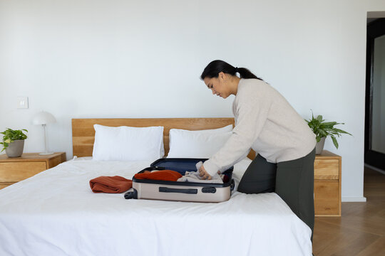 Asian woman kneeling on bed packing hard-shell suitcase in bedroom wearing beige sweater