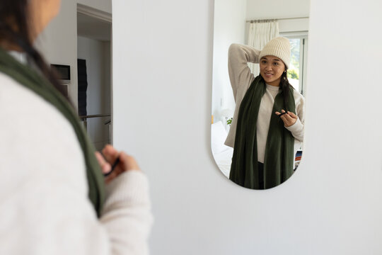 Asian woman adjusting beanie, green scarf while holding phone at oval mirror in bedroom, copy space