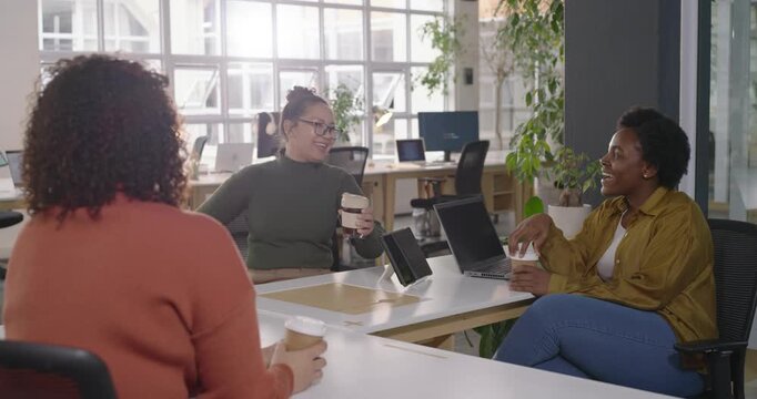 Female staff catching up as newcomer entering holding takeout cup at desk with laptop