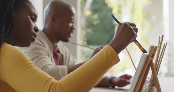 African American couple painting, man leaning, guiding, woman in yellow top adding paint to easel