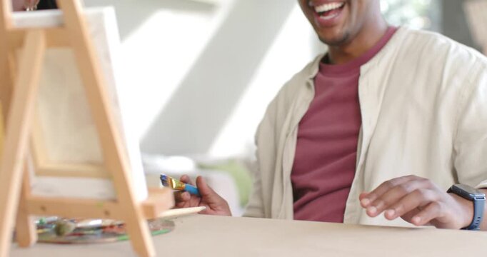 Two adults smiling, man picking paintbrush and guiding woman at easel at home, critiquing canvas