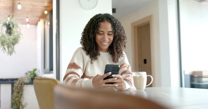 African American woman sitting at kitchen table, scrolling and replying to messages on smartphone