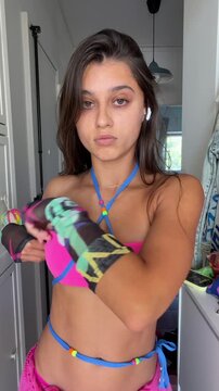 Young woman adjusts pink bandeau and blue string harness while pulling hair up in doorway, facing camera in summer styling moment