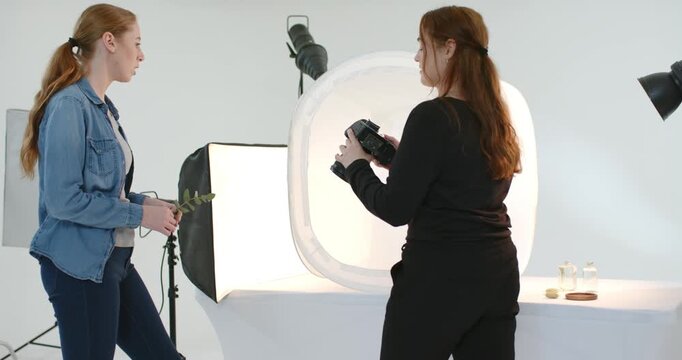 Female team holding sprig urging photographer adjusting DSLR placing props in light tent for sale