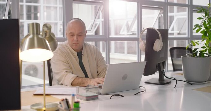 Man typing on laptop in office, working while camera moving closer revealing headphones on monitor