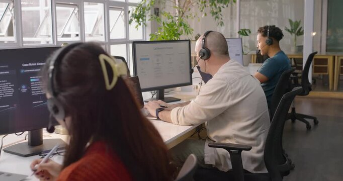 Camera panning, showing middle adult male typing laptop at open-plan bench, handling headset calls