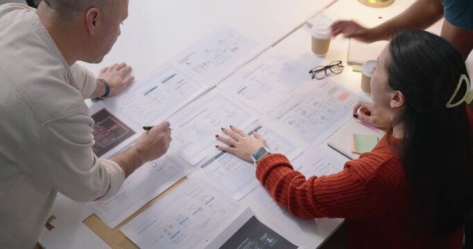 Diverse colleagues reviewing plans at office table, standing man approaching handing sticky notes