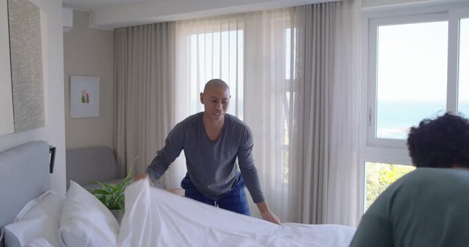 African American men making bed in coastal bedroom, bald man pulling duvet preparing for morning
