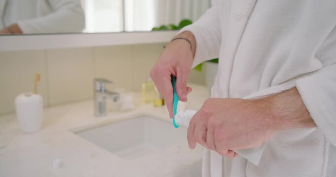 Man wearing robe removing tube cap, squeezing paste on teal brush, wetting, brushing teeth at sink