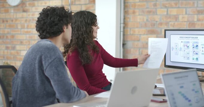 Diverse coworkers woman presenting printouts and pointing to monitor in office, reviewing concept