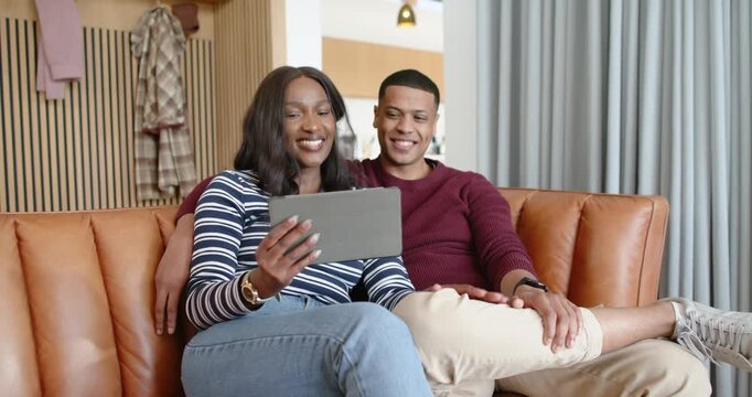 African American couple sitting on leather sofa, watching tablet call, connecting online and waving