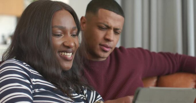 African American couple sitting on sofa, woman tapping tablet, watching funny clip and laughing