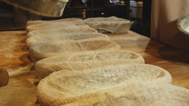 Baker pours wheat flour into proofing baskets with gauze sheets on wooden table closeup. Employee prepares bannetons for dough resting in bakery shop