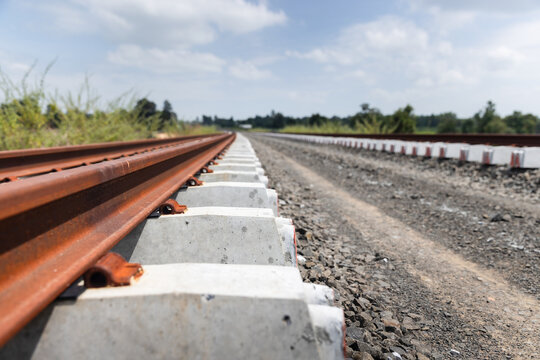 Low-angle view of steel railway rails mounted on concrete sleepers beside a gravel track bed, extending toward the horizon beneath a cloudy sky. Railway infrastructure and land transportation concept.