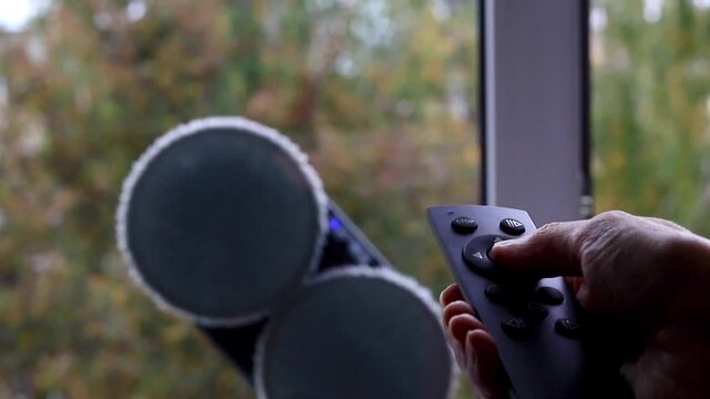 Person holding a remote control operates a robotic washer on the outdoor glass pane from a side view, managing automated cleaning and wiping of the exterior surface outside