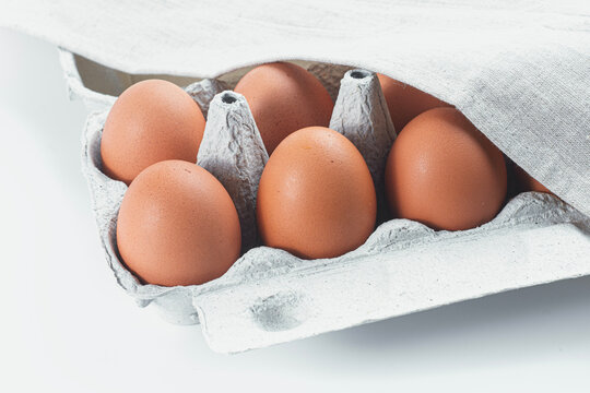 Brown chicken eggs in paper carton under white linen cloth on white background. Fresh organic farm product, healthy food and grocery concept.