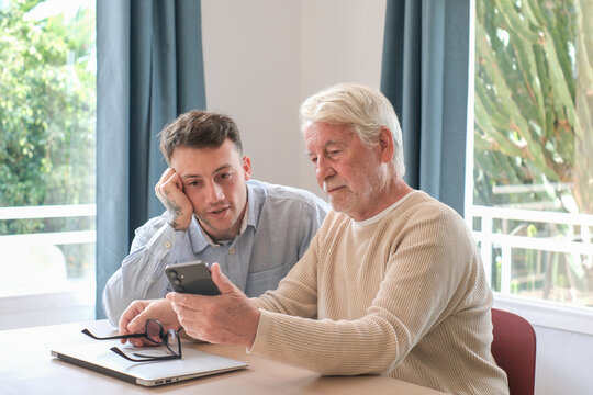 Young man teaching senior adult about smartphone features, connecting generations, sharing digital literacy skills, and navigating online technology together at home