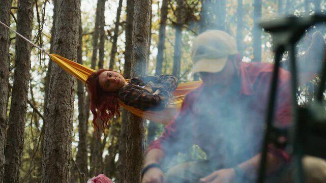 Red-haired woman talks from a hammock while a man tends a smoky campfire during camping in an autumn pine forest.