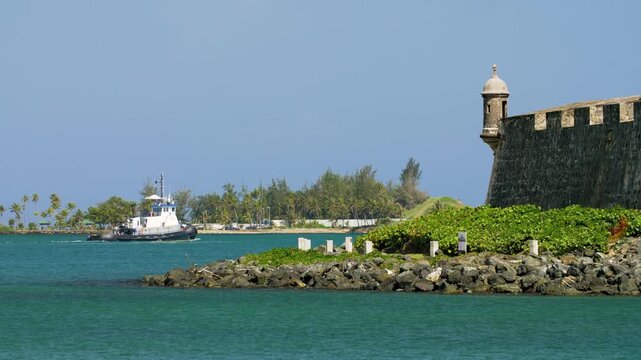 Tugboat travels along the San Juan bay as it departs into open sea and passes by the wall of the historic El Morro castle wall with Guard tower "Garita".