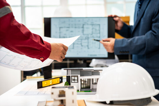 Architects review house plans beside a scale model, hard hat and computer screen in a bright modern office.