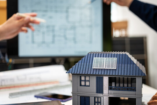 Miniature house with rooftop solar panels on architectural plans, with architects reviewing a home design in the background.