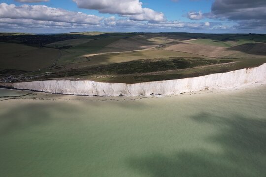 Aerial view of the iconic white chalk cliffs of the Seven Sisters coastline meeting the sea, with rolling green hills and a bright blue sky above in Seven Sisters, United Kingdom.