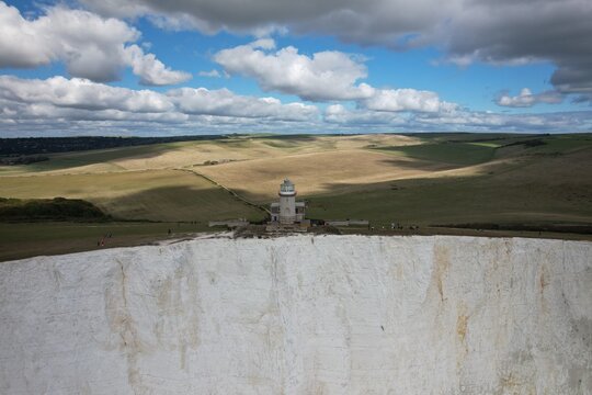 Aerial view of Belle Tout Lighthouse on white chalk cliffs, with rolling green hills and a cloudy sky in the background Seven Sisters, Wales, United Kingdom.
