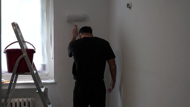 Back View of Painter Applying White Paint to Wall with Roller Tool in Bright Room Near Window Home Renovation DIY Project Concept Residential Interior
