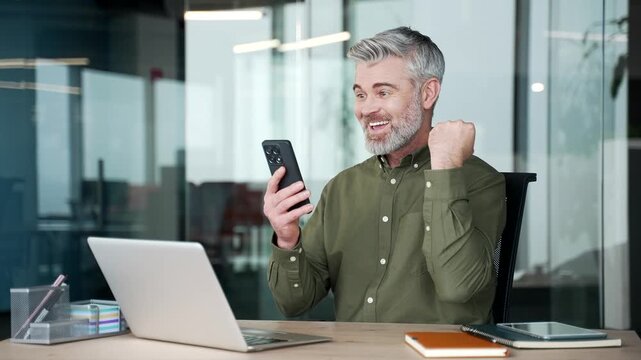 Happy gray-haired businessman celebrating success after receiving great news on smartphone at workplace. Smiling man reading positive message in office, expressing joy and excitement after achievement