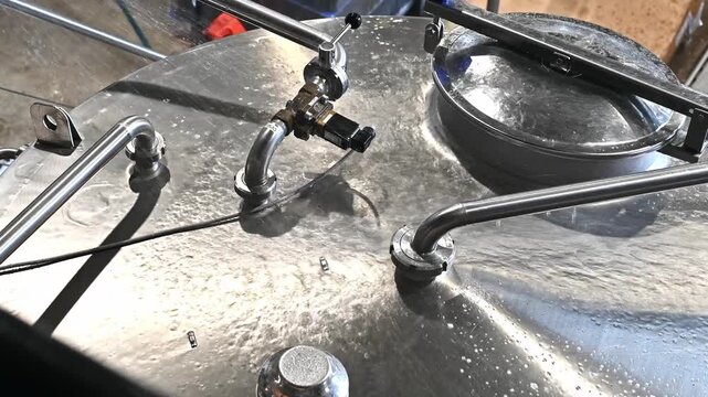 A high-angle close-up shot of water spraying and washing the stainless steel top hatch and pipes of a brewery or industrial processing tank.