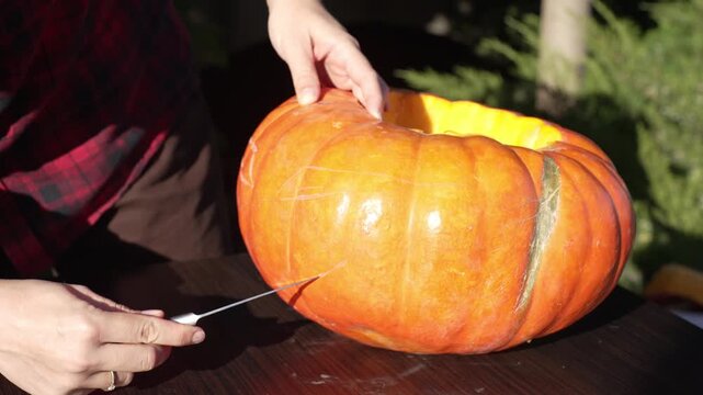 Woman scoring and tracing sinister smile outline on pumpkin with knife tip at sunset, static 4k 60fps slow motion footage