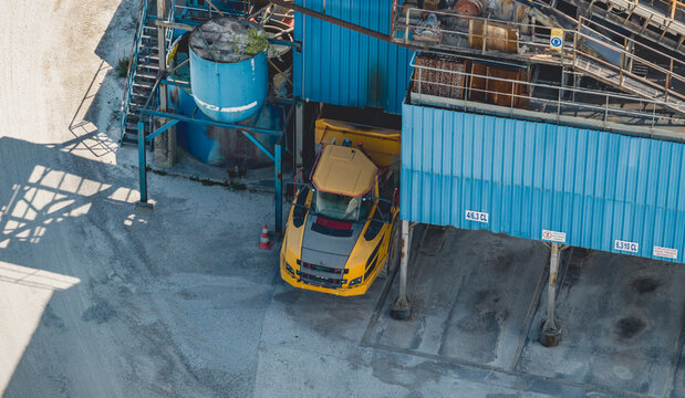 Aerial view of a yellow industrial dump truck parked beneath an aggregate processing facility with blue metal structures and gravel ground, Loriol-sur-Drome, Auvergne-Rhone-Alpes, France.