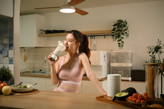 Young Asian Woman Drinking Protein Shake from Electric Shaker Bottle in Modern Kitchen with Health Supplements and Fresh Vegetables