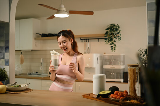 Young Asian Woman in Fitness Wear Holding Electric Shaker Bottle with Protein Supplement in Modern Kitchen